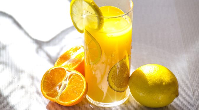 prim plan cu un pahar de limonada pe marginea caruia e o lamaie si langa se afla o lamaie intreaga in partea dreapta si o portocala taiata pe jumatate in partea stand - reprezentare a Vitaminei C in limonada